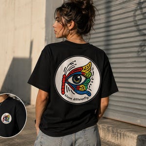 Può includere: T-shirt nero con grafica circolare sul retro raffigurante un occhio colorato e un'ala di farfalla. Il testo "bloom differently" è sotto la grafica. La maglietta è uno stile casual a maniche corte.