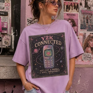 Può includere: Una t-shirt viola chiaro con una stampa grafica di un telefono cellulare vintage con un cuore sullo schermo, circondato da pianeti e stelle. Il testo recita Y2K CONNECTED e NOT JUST A SIGNAL, A SOUL FREQUENCY in una cornice rettangolare.