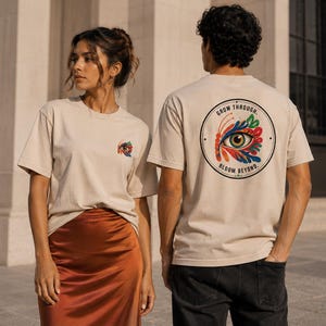 Può includere: Due persone indossano magliette beige. Una ha un colorato disegno di occhio e farfalla sul retro con la scritta "GROW THROUGH. BLOOM BEYOND." L'altra ha una versione più piccola del disegno sul davanti.