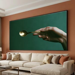 May include: A large rectangular wall art piece featuring a realistic depiction of a hand holding a metallic gold spoon against a solid dark green background. The artwork is framed in a thin black border and displayed above a beige sectional sofa.