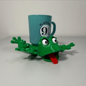 May include: A teal mug with the Platform 9 3/4 logo rests on a green frog-shaped holder. The frog has a red tongue and white eyes. The mug has a handle and is cylindrical.
