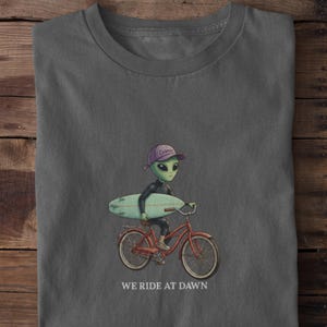 May include: Gray t-shirt featuring an alien riding a red bicycle and holding a surfboard. The alien wears a purple cap with the word "Cosmic" and a black suit. The text "WE RIDE AT DAWN" is printed at the bottom.