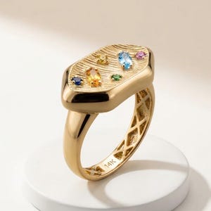 May include: A gold ring featuring a wide, textured, irregular-shaped top surface inlaid with six small, multi-coloured gemstones in various cuts. The inner band is marked with 14K, and the shank features an open, lattice-like geometric pattern.