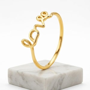 May include: A polished gold ring featuring the word love written in a continuous cursive script. The ring has a thin, smooth band and a bright, reflective metallic finish, showcasing the letters as the focal point of the jewelry piece.