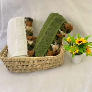 Peut inclure: Une serviette blanche et une serviette vert olive, toutes deux ornées de pompons décoratifs dans des tons marron, beige et vert sur les bords. Les serviettes sont pliées et placées dans un panier tressé à côté d'un petit pot blanc à fleurs jaunes.