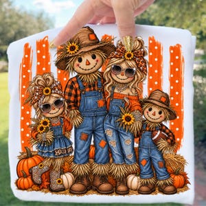 Op de afbeelding: Een wit t-shirt met een afbeelding van vier vogelverschrikkers in spijkeroveralls, geruite overhemden en hoeden. Het ontwerp bevat zonnebloemen, pompoenen en een football, tegen een achtergrond van oranje verticale strepen met witte stippen.