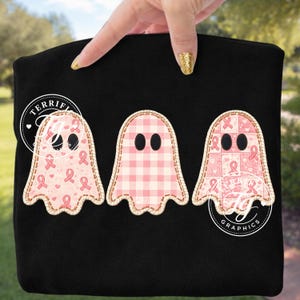Pode incluir: Um artigo de tecido preto com três formas de fantasmas bordadas. O primeiro fantasma tem um padrão de laços cor-de-rosa de consciencialização do cancro da mama, o segundo um padrão de xadrez vichy rosa e o terceiro um design patchwork rosa.