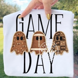 Puede incluir: Una camiseta blanca con el texto negro GAME DAY y tres apliques en forma de fantasma. El fantasma de la izquierda tiene un patrón de balones de fútbol americano y estrellas, el central parece un balón y el de la derecha tiene un patrón de cuadros.