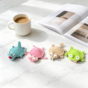 May include: Four small, round, crocheted animal figures in teal, pink, beige, and lime green. The teal figure has a red mouth, the pink one has two white eyes, the beige one has a small tail, and the lime green one has a pink tongue sticking out.