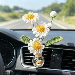 May include: A car air freshener featuring a small glass bottle with a teddy bear inside, topped with a cork and a bouquet of crocheted daisies. The daisies have white petals and yellow centers, with green leaves.