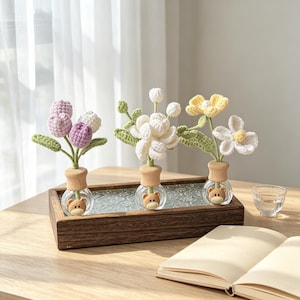 May include: Three decorative crochet flower arrangements in glass bottles with wooden stoppers and bear faces. The flowers are in shades of purple, white, and yellow with green leaves. The bottles sit in a wooden tray on a wooden table.