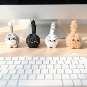 May include: Four small, round, crocheted cat figurines standing in a row. The cats are grey and white, solid black with green eyes, white with grey stripes, and fuzzy tan. Each has a tall, upright tail, embroidered facial features, and whiskers.