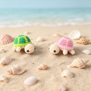 May include: Two small crocheted turtle figurines, one with a green and lime shell and the other with a pink and light pink shell. Both turtles have cream-colored heads, limbs, and black bead eyes, positioned on a sandy surface with seashells.