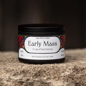 May include: A black candle tin with the text "Early Mass" and "A cup of Earl Grey tea." The label has a colorful floral design. The candle is on a stone surface.