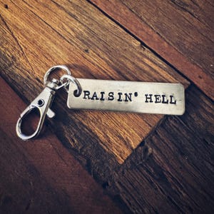May include: A silver keychain with the phrase "RAISIN' HELL" stamped on a rectangular metal tag. The keychain includes a silver clasp and a ring. The background is a wooden surface.