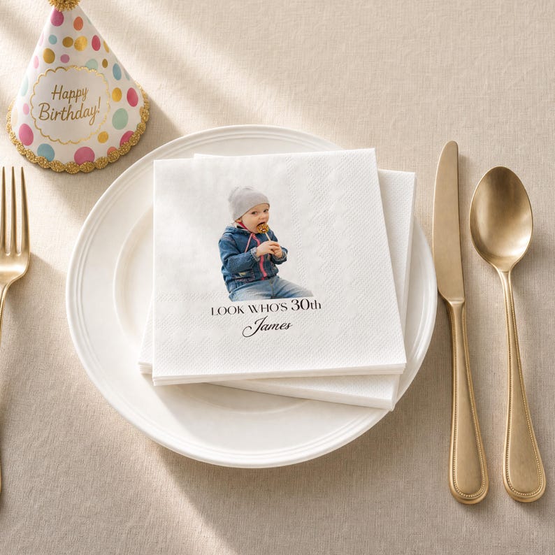 Puede incluir: Servilletas de papel blancas con una foto de un beb&eacute; con una piruleta y el texto "LOOK WHOS 30th James". Un tenedor, un cuchillo y una cuchara dorados est&aacute;n sobre un plato blanco. Un gorro de fiesta con las palabras "Happy Birthday!" tambi&eacute;n est&aacute; en la imagen.