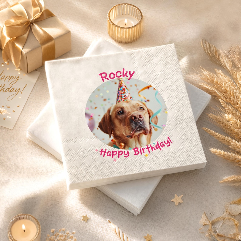 Puede incluir: Servilletas cuadradas blancas con un patr&oacute;n de diamantes. Una imagen circular de un perro con un gorro de fiesta est&aacute; impresa en la servilleta superior, con el nombre "Rocky" y las palabras "Happy Birthday!" en rosa. Una caja de regalo y una vela est&aacute;n en el fondo.