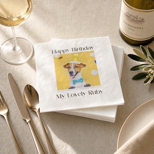 Puede incluir: Servilletas de papel blancas con una imagen impresa de un perro con gorro de fiesta y pajarita. El texto "Happy Birthday" y "My Lovely Ruby" est&aacute; impreso. Un vaso de vino blanco y una botella de Chardonnay est&aacute;n en el fondo.