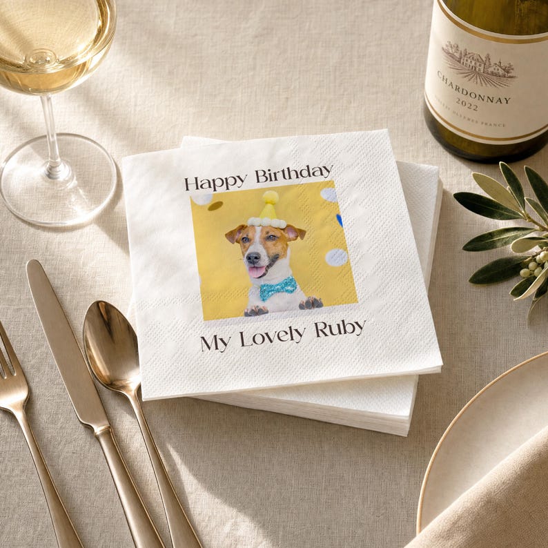 Puede incluir: Servilletas de papel blancas con una imagen impresa de un perro con gorro de fiesta y pajarita. El texto "Happy Birthday" y "My Lovely Ruby" est&aacute; impreso. Un vaso de vino blanco y una botella de Chardonnay est&aacute;n en el fondo.