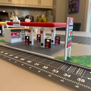 May include: A miniature model of a 7-Eleven gas station with a red and white canopy, a sign displaying "1.10", and a Canadian flag. The model sits on a gray surface with a black ruler.
