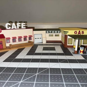 May include: A model of a cafe, store, and petrol station. The cafe features a red awning and the word "CAFE". The store has the word "STORE". The petrol station has the word "GAS" and two petrol pumps.