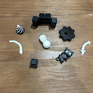 May include: A collection of small, 3D-printed objects in black and white, including a cat, a geometric shape, and a spiral. The objects are arranged on a wooden surface.
