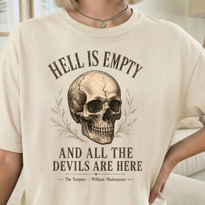 May include: Cream-colored t-shirt featuring a detailed skull graphic with the text "HELL IS EMPTY AND ALL THE DEVILS ARE HERE" above and below the skull. The quote is from William Shakespeare's "The Tempest".