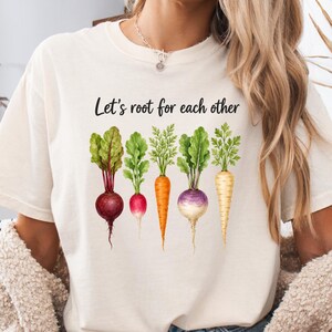 Può includere: Una t-shirt a maniche corte color crema con una grafica di cinque colorati ortaggi a radice, tra cui una barbabietola, un ravanello, una carota, una rapa e una pastinaca. Sopra le verdure, il testo recita Let's root for each other in corsivo nero.