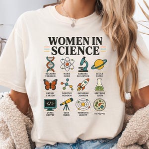 May include: A cream-coloured t-shirt featuring the text "WOMEN IN SCIENCE" in black, accompanied by twelve colourful scientific icons and the names of notable figures including Rosalind Franklin, Marie Curie, and others printed in a grid layout.