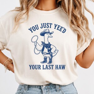 May include: A cream-colored short-sleeve t-shirt featuring a blue graphic of a goose dressed as a cowboy with a hat, bandana, vest, and boots, holding a lasso. The text above and below the goose reads, "YOU JUST YEE'D YOUR LAST HAW."