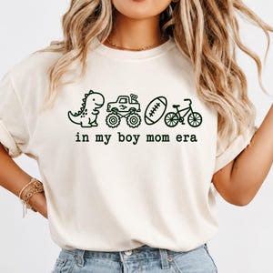 May include: A cream-coloured short-sleeve t-shirt featuring a dark green graphic print of a dinosaur, a monster truck, a football, and a bicycle. Below the icons, the text reads in a typewriter-style font: in my boy mom era.