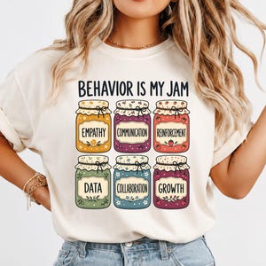 Peut inclure: Un t-shirt couleur crème avec le texte BEHAVIOR IS MY JAM au-dessus de six illustrations colorées de bocaux en verre. Chaque bocal porte une étiquette avec un mot : EMPATHY, COMMUNICATION, REINFORCEMENT, DATA, COLLABORATION et GROWTH, avec des motifs floraux.
