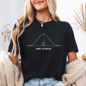 May include: A black short-sleeved t-shirt featuring a white graphic of a bell curve with a small ghost hanging from the peak. Below the graphic, the text reads NORMAL DISTRIBUTION in a clean, sans-serif font. The shirt has a crew neckline.