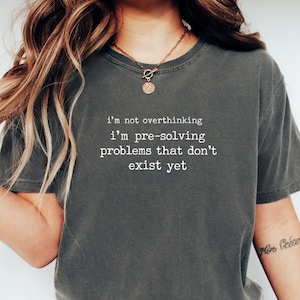 Könnte beinhalten: Ein anthrazitfarbenes Kurzarm-T-Shirt mit weißem Text im Schreibmaschinenstil: i'm not overthinking i'm pre-solving problems that don't exist yet. Das T-Shirt hat einen Rundhalsausschnitt und eine lockere Passform aus weichem, verwaschenem Stoff.