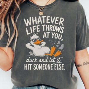 Może przedstawiać: Ciemnoszara koszulka z napisem "WHATEVER LIFE THROWS AT YOU, duck and let it HIT SOMEONE ELSE." Na koszulce znajduje się postać kaczki z kreskówki w okularach przeciwsłonecznych. Koszulka to element odzieży codziennej.