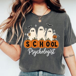May include: A charcoal gray t-shirt featuring a graphic of three ghosts behind five orange pumpkins. The ghosts hold a mug, make a peace sign, and carry a jack-o'-lantern bucket. The text reads "SCHOOL" on the pumpkins and "Psychologist" below.