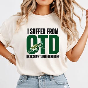 May include: A cream-coloured short-sleeve t-shirt featuring the black text I SUFFER FROM and the green text OTD with a graphic of a green sea turtle replacing the letter T. Below the graphic is the black text OBSESSIVE TURTLE DISORDER.