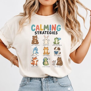 Puede incluir: Una camiseta de color crema con el texto "CALMING STRATEGIES" en letras coloridas sobre nueve ilustraciones de animales realizando diversas actividades, incluyendo un oso, conejo, rana, pájaro, gato, perro, zorro, tortuga y ratón con etiquetas.