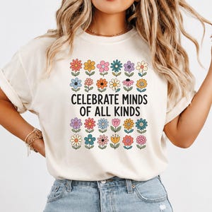 Puede incluir: Una camiseta de manga corta de color crema con un estampado gráfico de flores coloridas dispuestas en filas sobre y debajo del texto negro que dice, Celebrate Minds of All Kinds. La camiseta se combina con unos vaqueros azul claro.