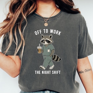 May include: A charcoal grey short-sleeve t-shirt featuring a graphic of a raccoon wearing sage green scrubs, holding a coffee cup, and walking. The text above the raccoon reads OFF TO WORK and the text below reads THE NIGHT SHIFT in white.