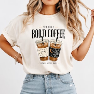 Peut inclure: Un t-shirt couleur crème avec un motif de deux tasses de café glacé et des pailles noires. Une tasse présente un fantôme et des étoiles, l'autre une lune et une toile d'araignée. Le texte indique FRESHLY BOO'D COFFEE et BREWED AFTER DARK.