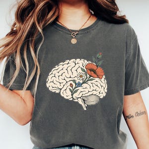 Puede incluir: Una camiseta de manga corta gris carbón con una ilustración gráfica de un cerebro humano con flores coloridas, incluyendo una amapola naranja grande, una margarita blanca y pequeñas flores azules que brotan de la parte superior.