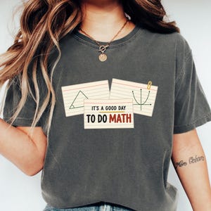 Può includere: Una t-shirt a maniche corte grigio antracite con una grafica raffigurante tre schede a righe. Le schede mostrano un triangolo, un grafico a parabola e il testo IT'S A GOOD DAY TO DO MATH in caratteri neri, arancioni e rossi con una graffetta dorata.