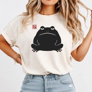 Puede incluir: Una camiseta de manga corta color crema con un gráfico grande en negro sólido de un sapo sentado con expresión gruñona. Sobre la cabeza del sapo, hay un pequeño icono cuadrado rojo que contiene un símbolo blanco estilizado de montaña.