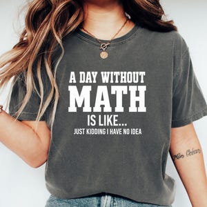 Puede incluir: Una camiseta de manga corta en color gris carbón con letras blancas en bloque que dicen A DAY WITHOUT MATH IS LIKE... JUST KIDDING I HAVE NO IDEA. La prenda tiene cuello redondo y un corte relajado, confeccionada en algodón.