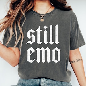 May include: A charcoal gray short-sleeve t-shirt featuring the text "still emo" printed in a white, distressed, gothic-style font across the chest. The garment has a crew neckline and a relaxed fit, made from a solid-colored cotton fabric.