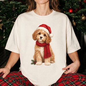 Peut inclure: Un t-shirt à manches courtes de couleur crème arborant une illustration graphique d'un chiot brun au pelage duveteux, portant un bonnet de Père Noël rouge et une écharpe en tricot rouge assortie, avec une branche de pin et des baies rouges.