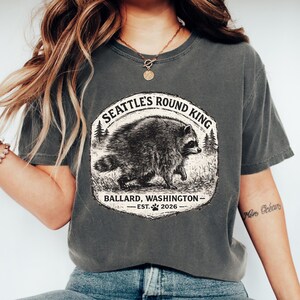 以下が含まれることがあります： チャコールグレーの半袖Tシャツ。森の中にいるアライグマのグラフィックが描かれています。テキストには「SEATTLE'S ROUND KING」「BALLARD, WASHINGTON」「EST. 2026」とあり、アライグマのイラストの下には小さな足跡のアイコンが配置されています。