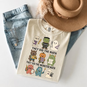 May include: A cream-coloured t-shirt featuring a graphic of nine cartoon monsters holding maths tools and supplies. The text reads "THEY DID THE MATH" and "THEY DID THE MONSTER MATH" in black capital letters. The shirt is paired with blue jeans.