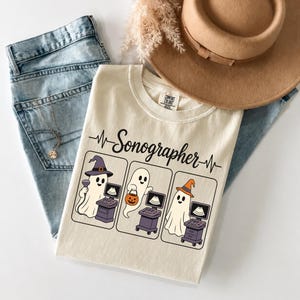 Puede incluir: Una camiseta color crema con el texto "Sonographer" sobre tres paneles rectangulares. Cada panel muestra un fantasma blanco con una máquina de ultrasonido. Los fantasmas llevan un sombrero de bruja morado, un cubo de calabaza o un sombrero naranja.